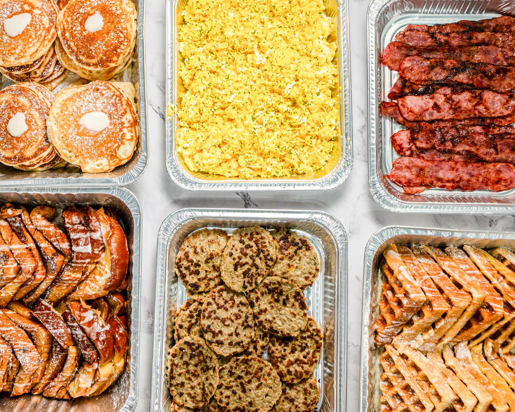 Six full catering trays: pancakes, scrambled eggs, beef bacon, French toast, turkey sausage, and waffles — laid out on a marble surface.